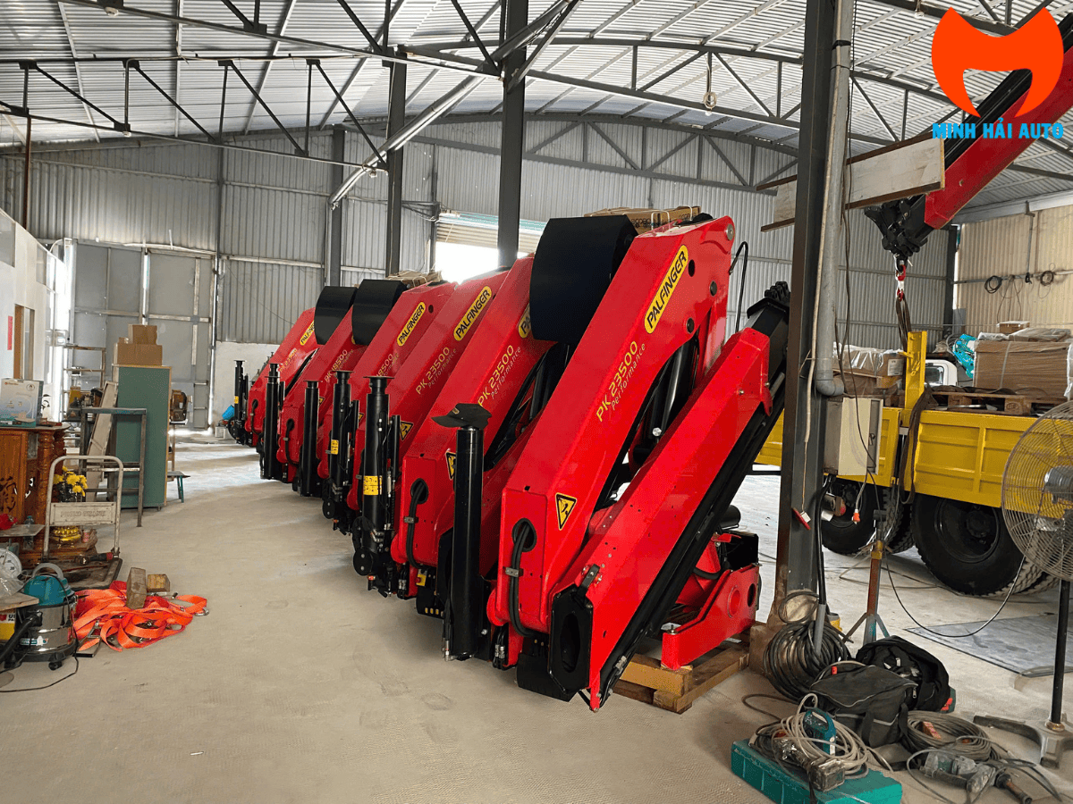 Cẩu gấp khúc Palfinger PK 8500 3 tấn màu cam được lắp đặt trên xe tải, đang vươn cần để nâng hàng tại một nhà xưởng.