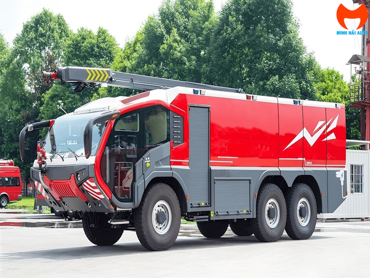 Xe cứu hỏa sân bay điện ARFF 6x6 màu đỏ đang phun một luồng nước mạnh mẽ trên đường băng, phía sau là một chiếc máy bay thương mại đang đỗ, thể hiện sự sẵn sàng và hiệu quả trong công tác PCCC tại sân bay.