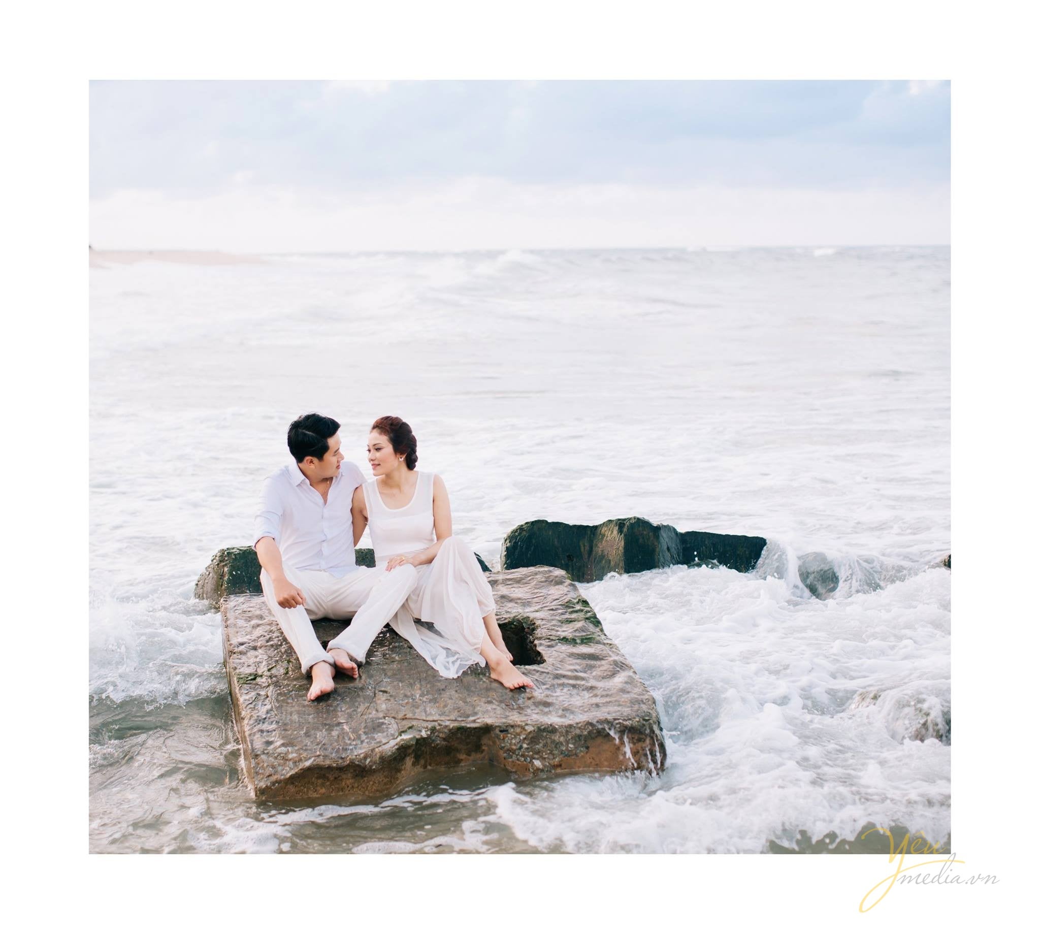cô dâu chú rể ngồi trên mỏm đá biển sóng đánh bọt trắng xóa