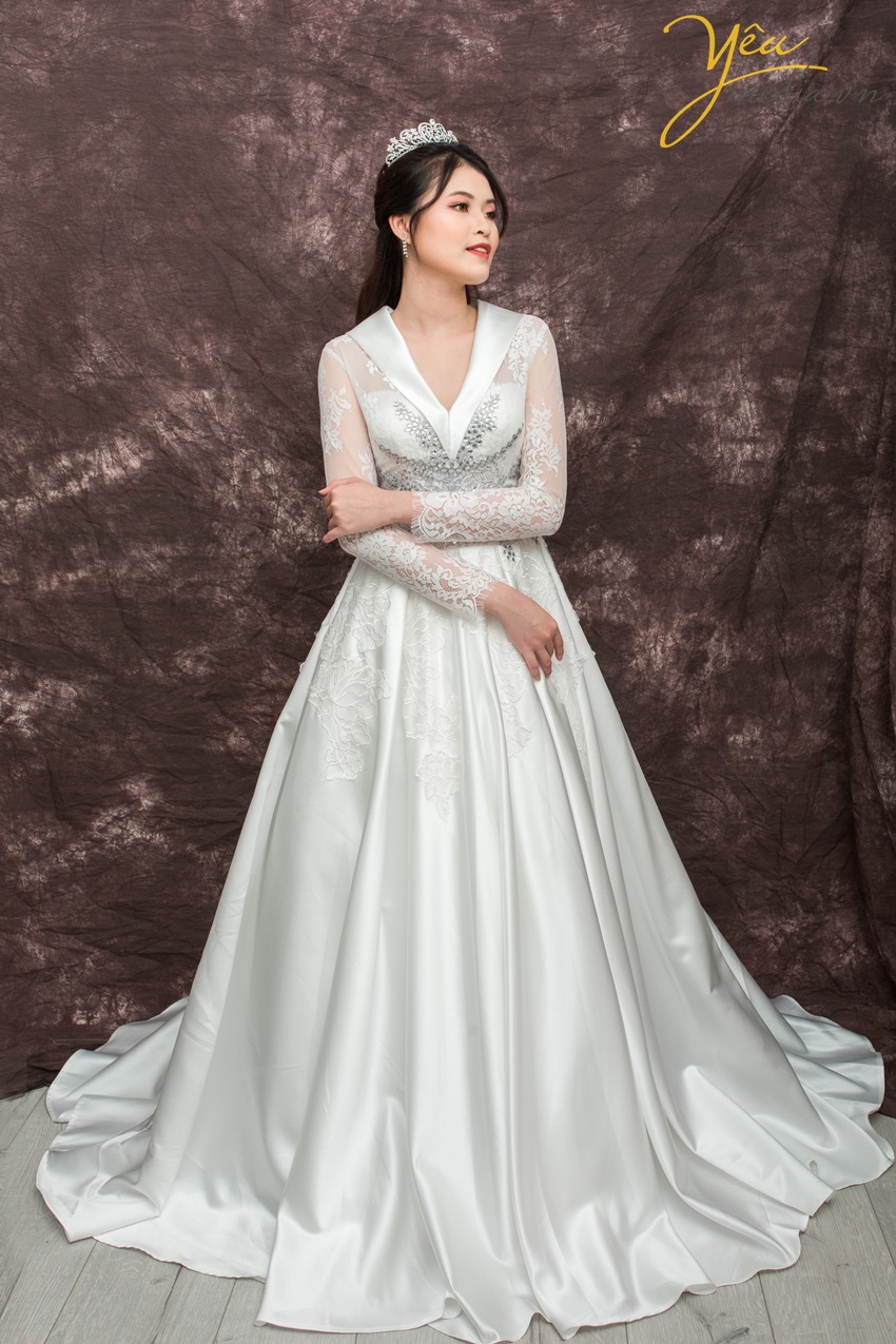 Lấy cảm hứng của hoàng gia, bộ váy cưới sang trọng tên Kate này đang được rất nhiều cô dâu ưa chuộng tại Yêu Media. Phong cách sang trọng với phần cổ áo độc đáo vắt chéo tam giác, phần đính hạt cườm nổi bật lấp lánh. Phần ren trên tay áo hài hòa và nhẹ nhàng, cổ tay dài, với phần ren tràn đính từ eo xuống thân váy phi bóng, càng tạo nên vẻ cổ điển cho cô dâu của Yêu Media.