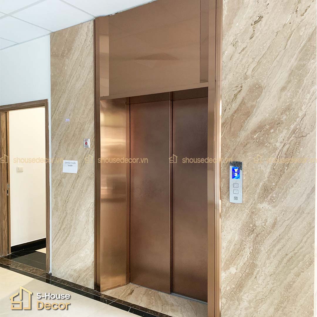 Khung bao cửa tầng bản rộng bằng inox vàng hồng xước, kết hợp decal trang trí hiện đại, tăng tính thẩm mỹ cho khu vực thang máy.