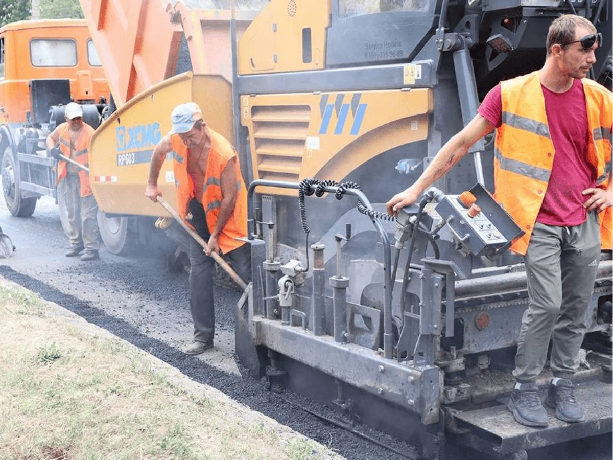 Máy rải thảm XCMG RP603 màu vàng đang rải nhựa đường, hai công nhân áo phản quang cầm bàn xoa san phẳng mép thảm, một người đứng trên bậc điều khiển