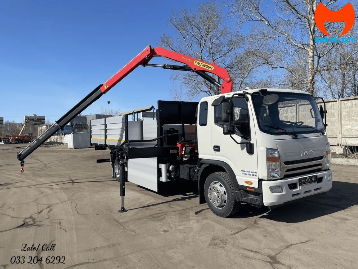 Xe tải JAC màu trắng gắn cẩu gập Palfinger đỏ, cần vươn ra và chân chống hạ xuống nền bê tông tại bãi xe