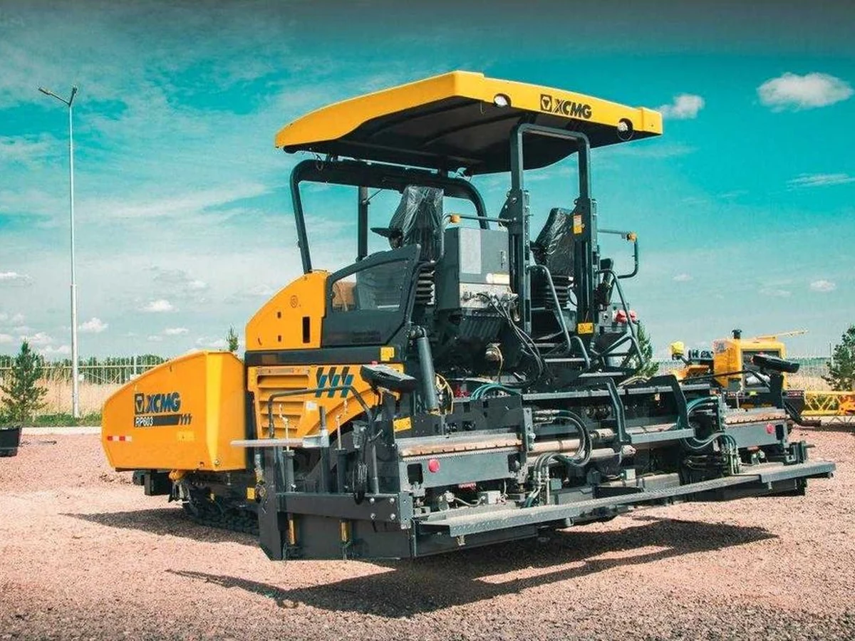 Máy rải thảm XCMG RP603 màu vàng đỗ tại bãi, chụp chếch trước bên trái, guồng thảm mở rộng hai bên và mái che buồng lái