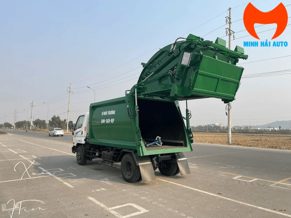 Xe cuốn ép rác thùng màu xanh đang nâng đuôi thùng để xả rác tại điểm tập kết