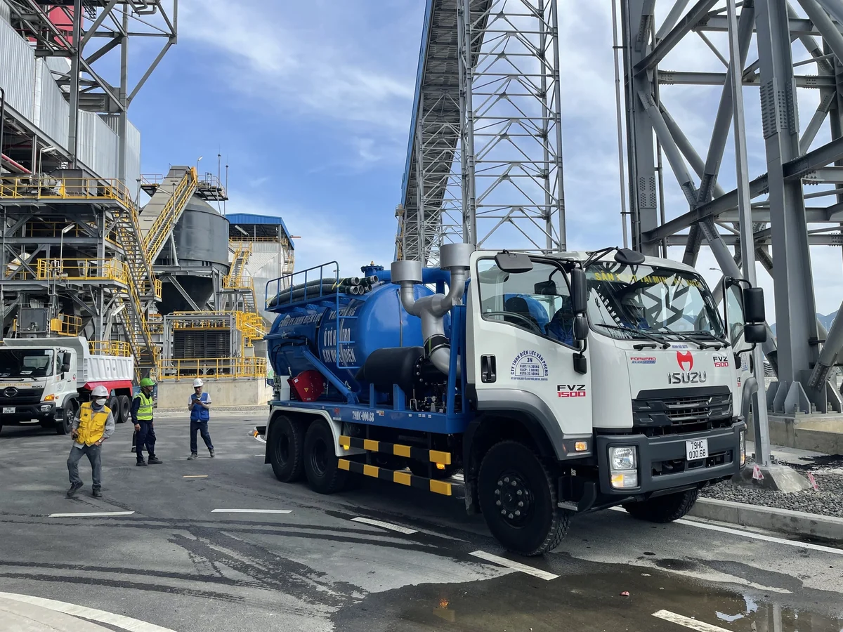 Xe hút tro bay công nghiệp đỗ trong khuôn viên nhà máy nhiệt điện, phía sau là hệ thống silo và kết cấu thép