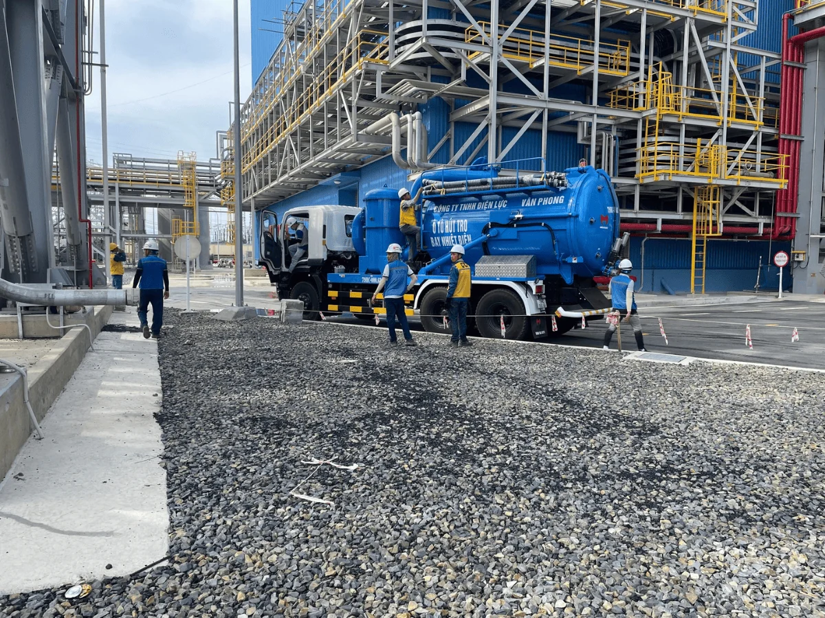 Nhân viên vận hành xe hút tro bay tại khu vực nhà máy nhiệt điện, xe đỗ cạnh kết cấu thép và đường ống