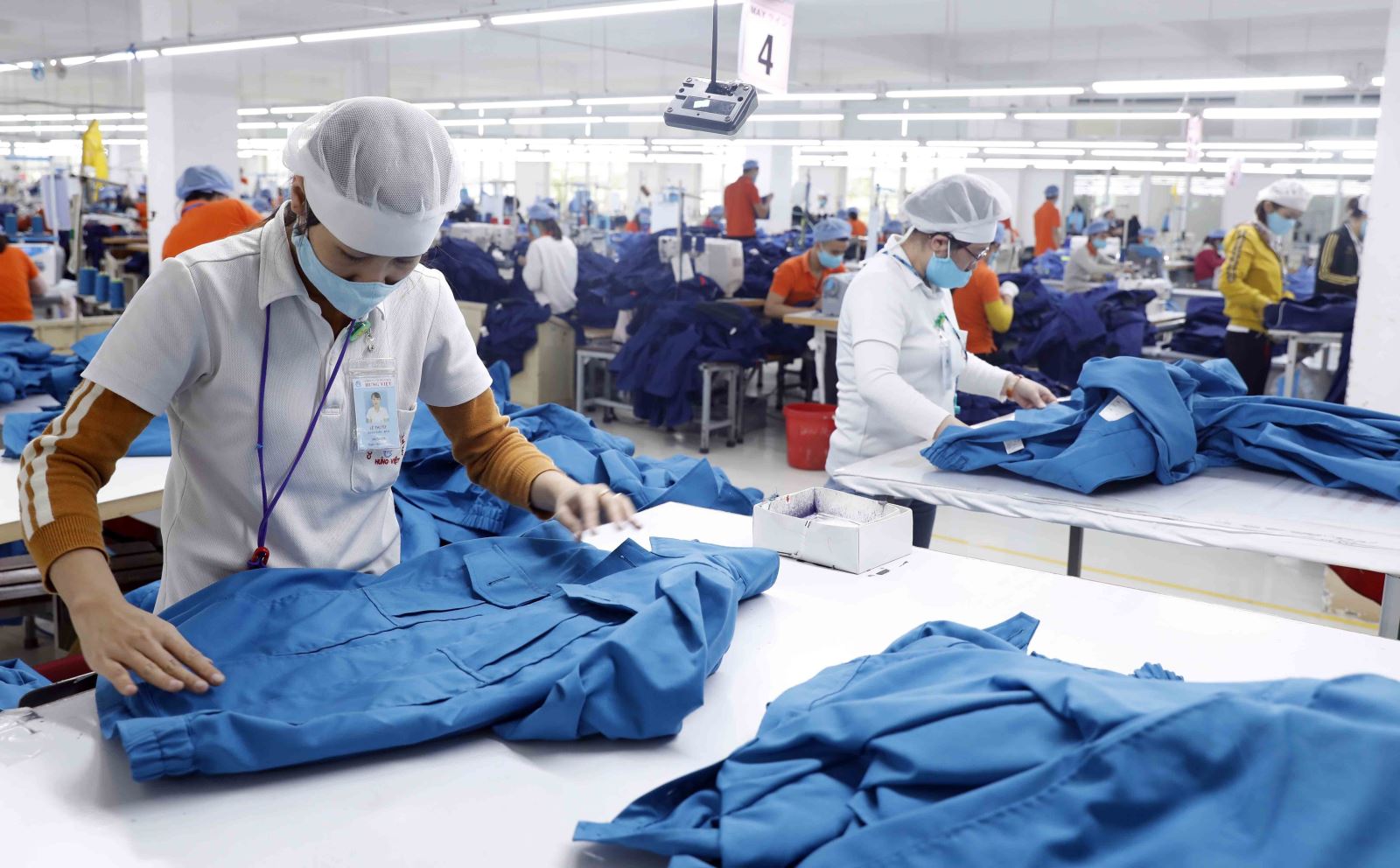 Công nhân mặc đồ bảo hộ làm việc tại xưởng may với các bàn máy và vải xanh