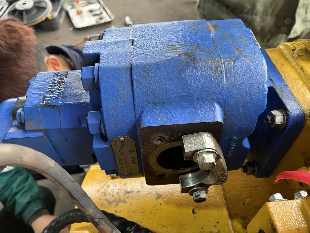 Cụm bơm thủy lực chính (bơm công tác/bơm lái kép) màu xanh được thợ kỹ thuật gá lắp hoàn chỉnh