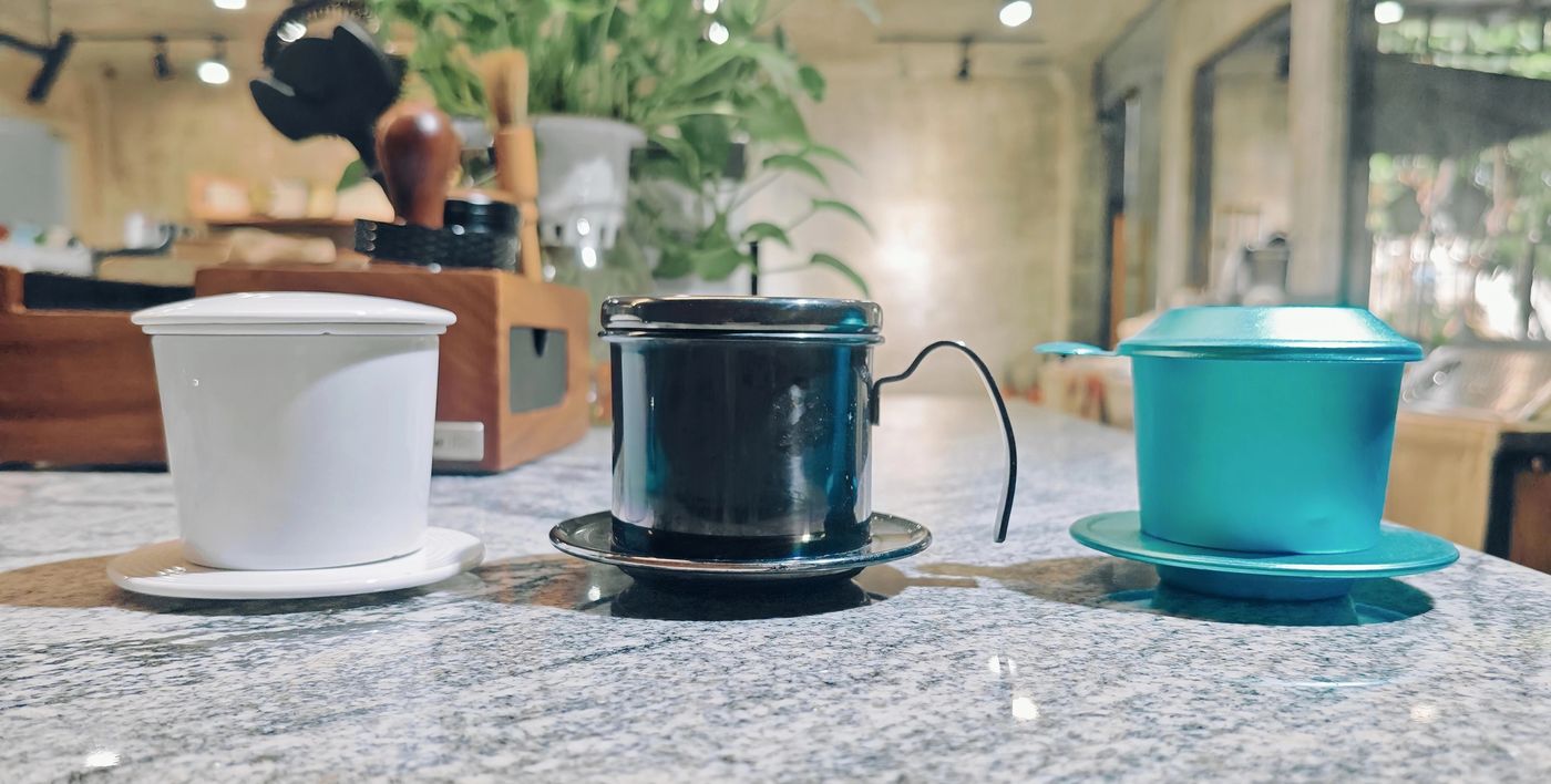 Ba loại phin cà phê Việt Nam đặt cạnh nhau tại showroom Volcano: phin sứ trắng, phin inox 304 đen nắp gài xoáy, phin nhôm anod xanh ngọc