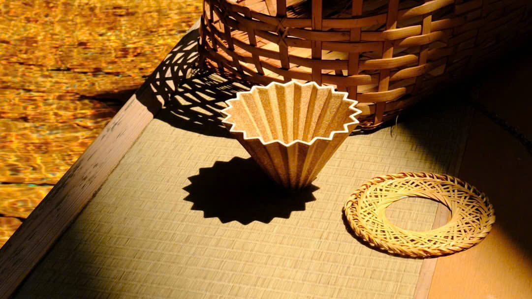 Phễu Origami Dripper S Rinko EC Limited đặt cạnh giỏ đan tre và đĩa tre thủ công dưới ánh sáng ấm