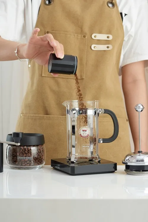 Bàn tay đổ bột cà phê vào bình French Press đặt trên cân điện tử