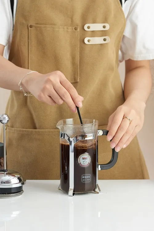 Bàn tay dùng muỗng khuấy nhẹ bột cà phê trong bình French Press