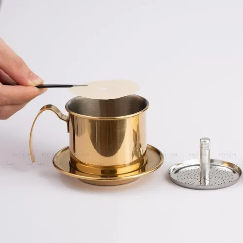 Bàn tay đặt tờ giấy lọc tròn vào lòng phin cà phê inox màu gold đặt trên đĩa lót