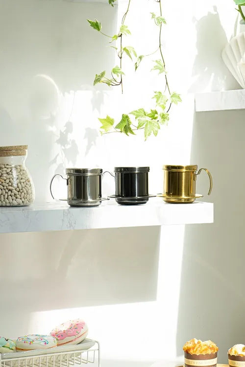 Ba phin cà phê inox màu bạc đen và gold bày trên kệ trắng cạnh cây xanh