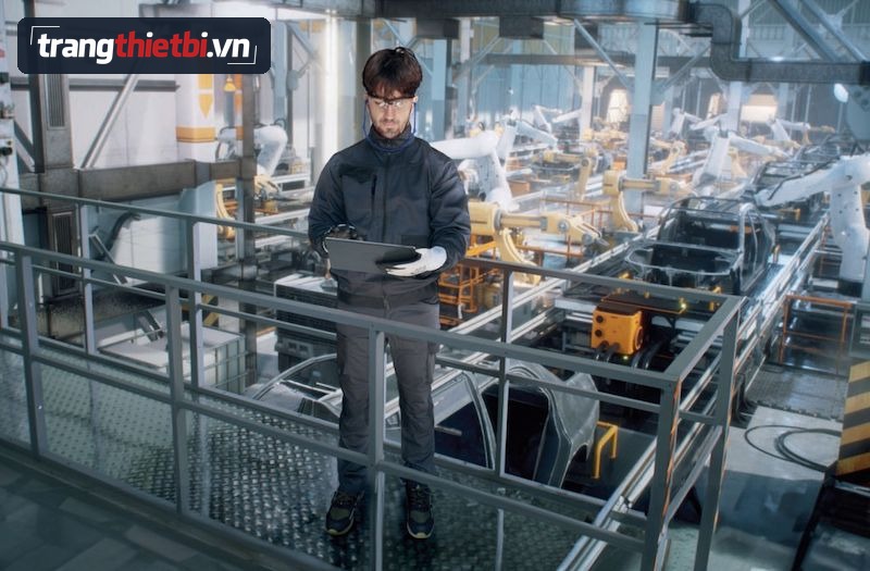 Kỹ sư đeo kính bảo hộ tai nghe chống ồn mang găng tay và giày bảo hộ đang cầm máy tính bảng giám sát dây chuyền robot lắp ráp ô tô