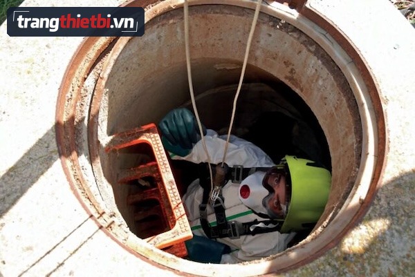 Công nhân đội mũ bảo hộ đeo mặt nạ phòng độc mặc áo chống hóa chất và dây đai an toàn đang leo xuống hầm cống hạn chế