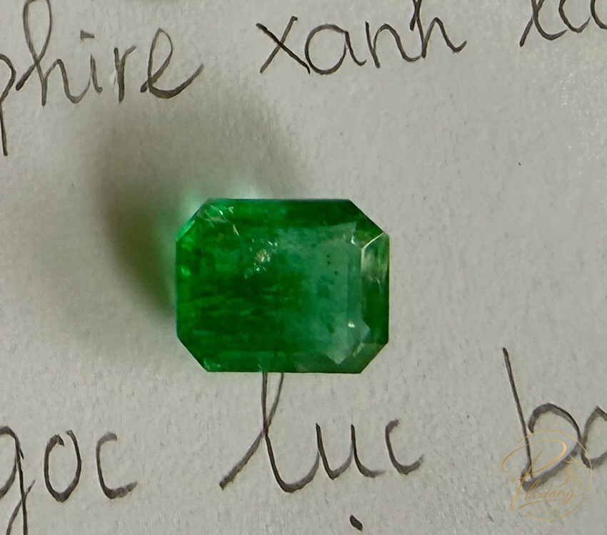 Viên ngọc lục bảo (Emerald) cắt giác emerald-cut màu xanh lục đậm, đặt cạnh dòng chữ viết tay ghi 'ngọc lục bảo' dùng để nhận diện loại đá