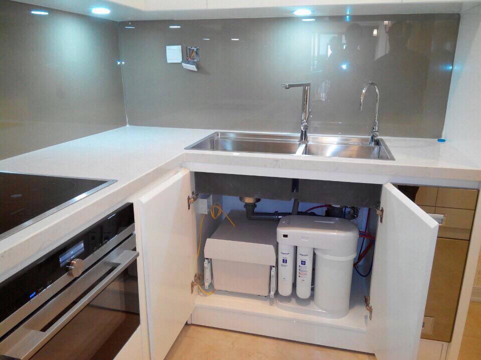 Máy lọc nước đặt bên phải khoang tủ bếp màu trắng, cạnh xi-phông và đường ống dưới chậu
