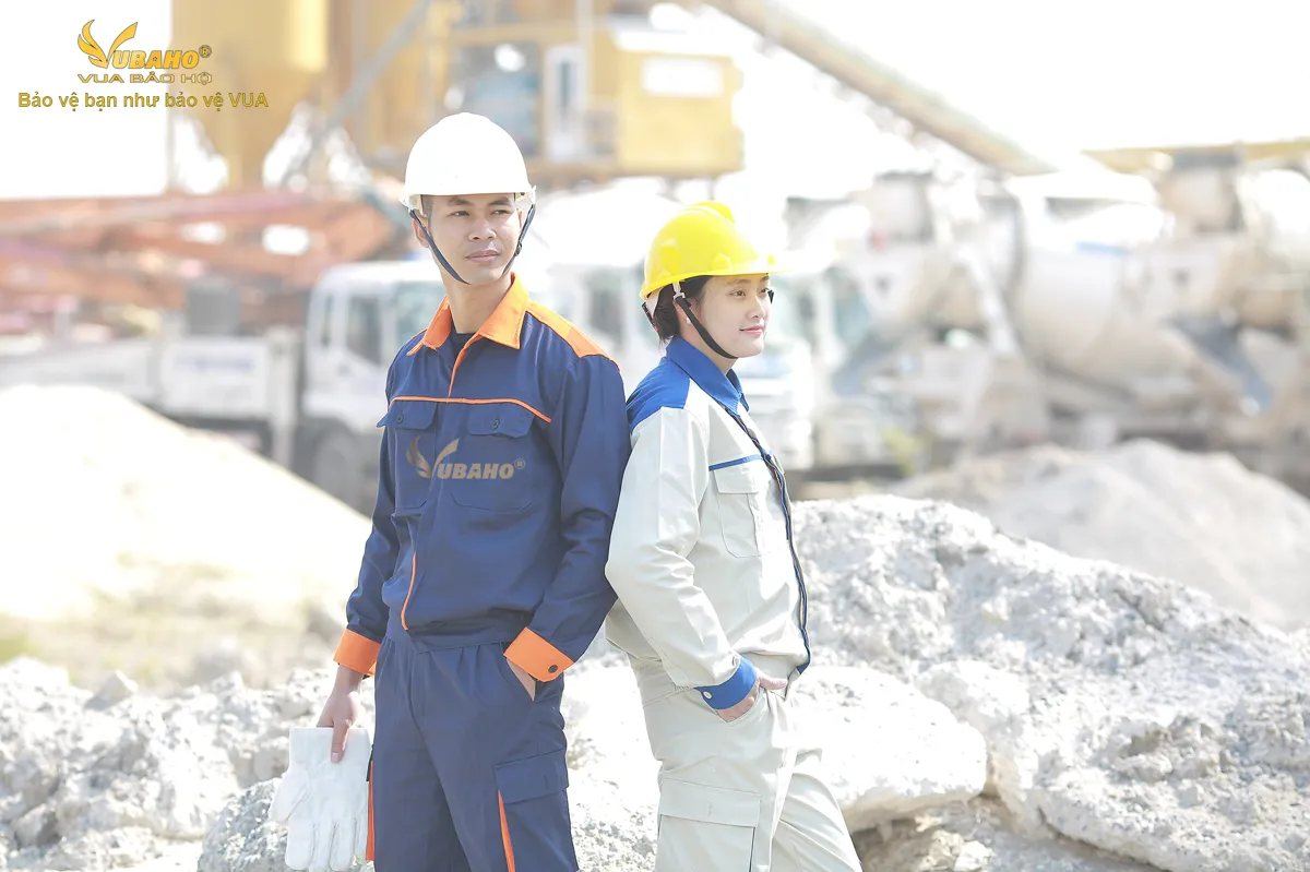 Hai công nhân mặc quần áo bảo hộ lao động tại công trường bê tông: bộ tím than phối cam và bộ ghi sáng phối xanh
