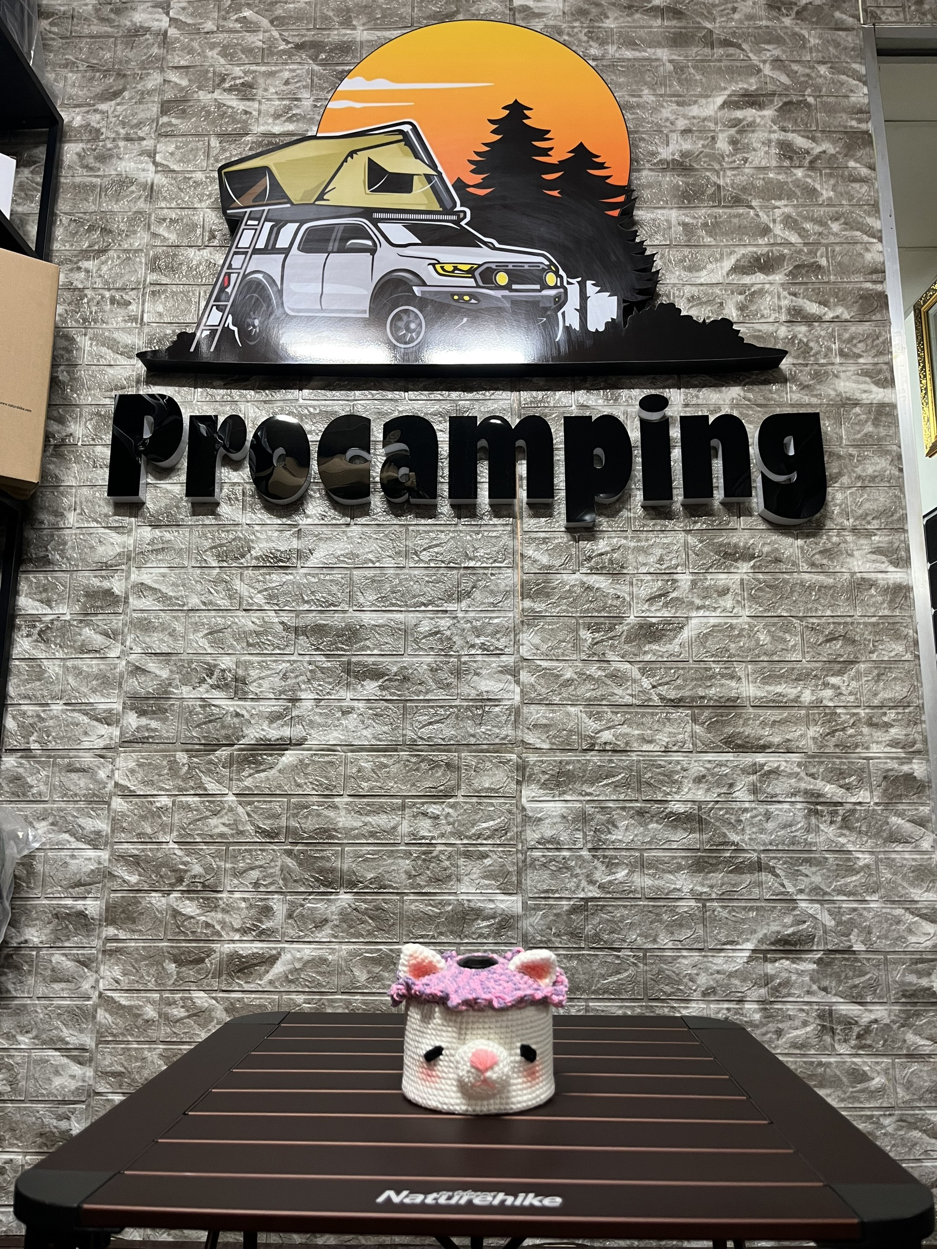 Áo len bọc bình gas mini hình mèo trắng đặt trên bàn, phía sau bảng hiệu Procamping