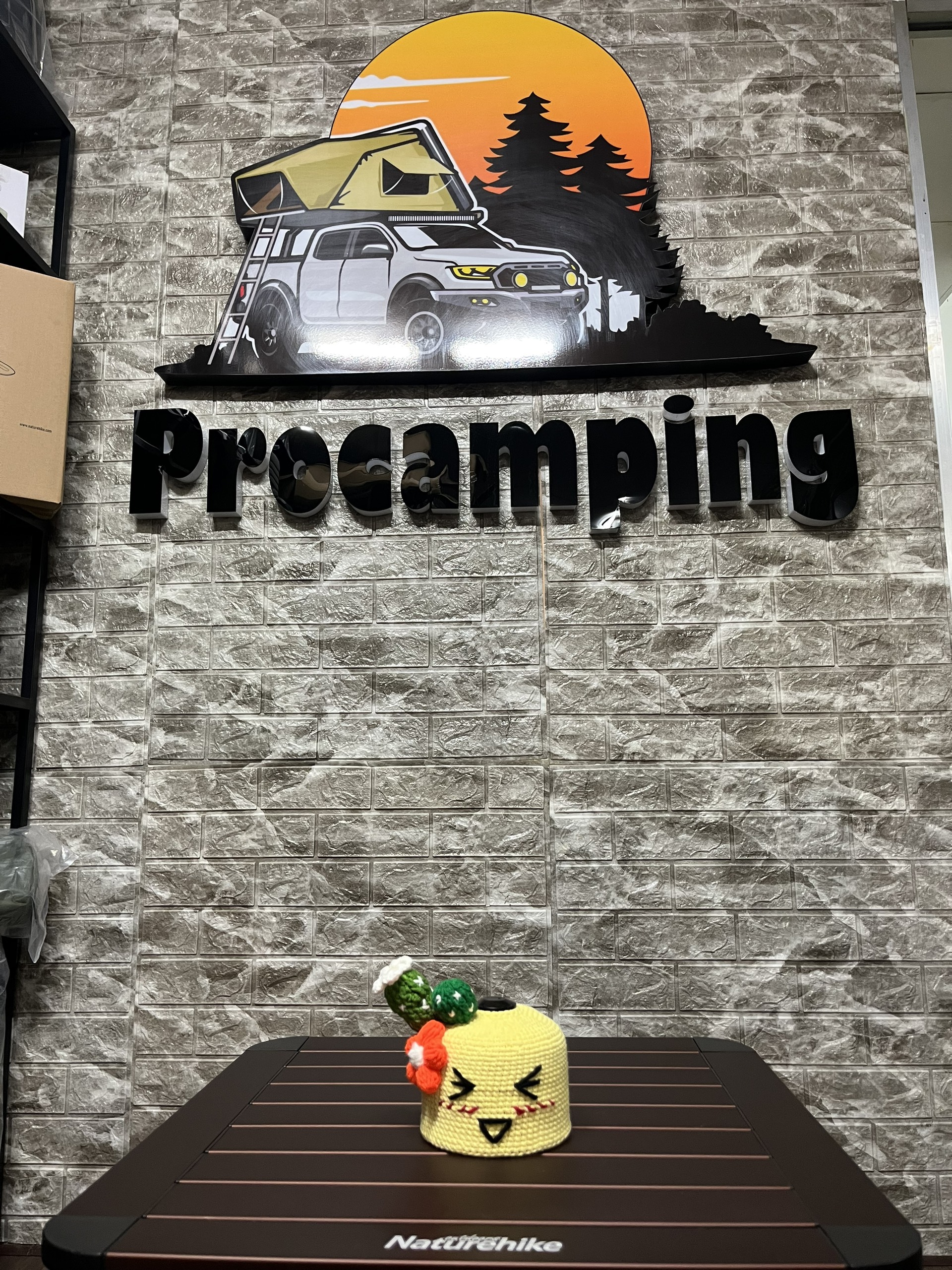 Áo len bọc bình gas mini hình mặt vàng cam đặt trên bàn, phía sau bảng hiệu Procamping
