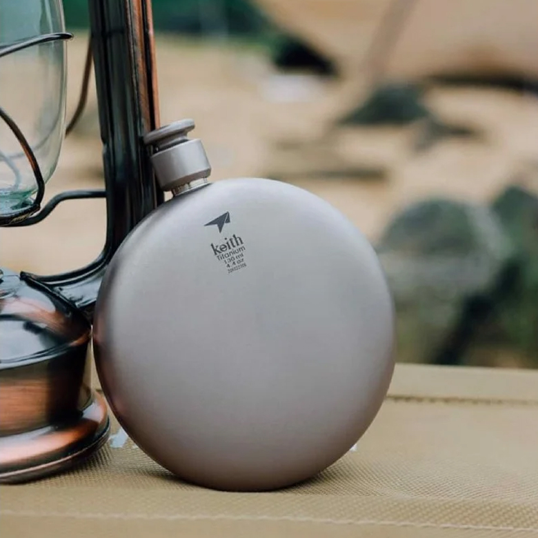 Keith Bình Rượu Titanium Round Flask 130ml màu xám đặt trên bàn gỗ cạnh đèn lồng cắm trại