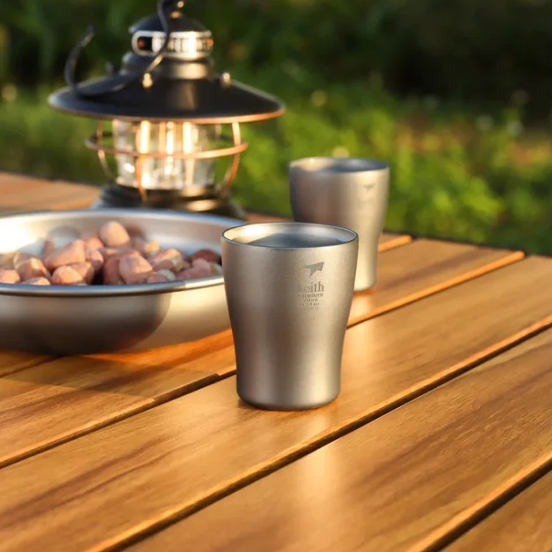 Ly Uống Rượu Keith Titanium Wine Cup 10ml trên bàn gỗ cạnh đĩa hạt và đèn lồng cắm trại