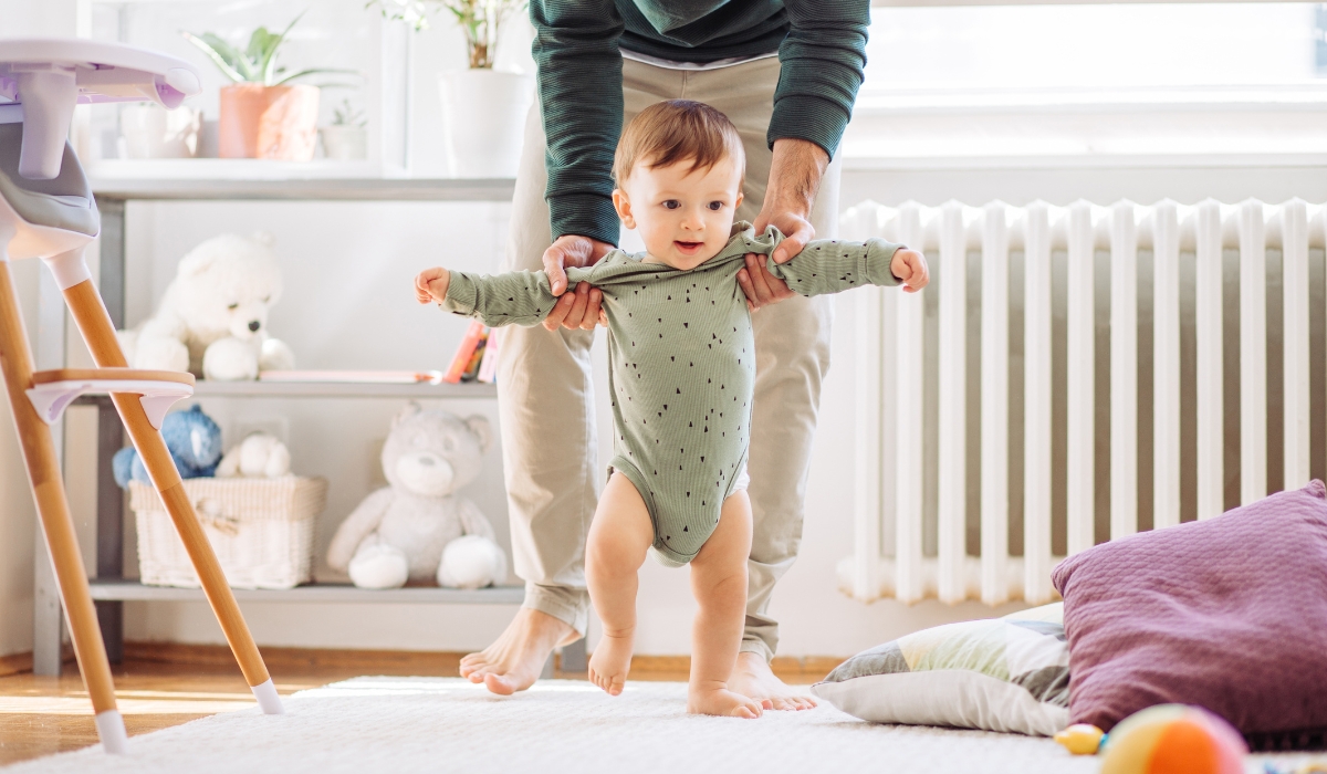 Trẻ nhỏ đang được cha hỗ trợ tập đi, với hai tay cha giữ vững, trong không gian sáng sủa và ấm áp
