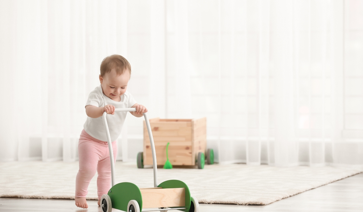 Trẻ nhỏ đang vui vẻ đẩy xe tập đi bằng gỗ trong không gian sáng sủa, thể hiện sự phát triển tự nhiên và an toàn trong quá trình học đi