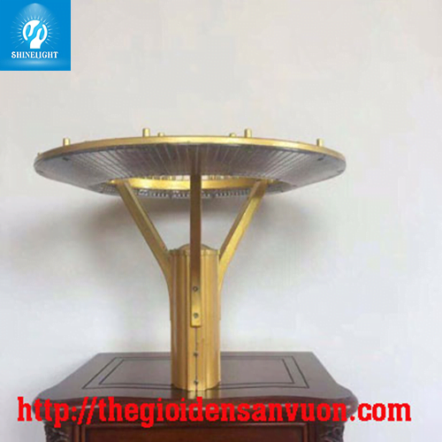 Đầu đèn sân vườn SLHD86 màu vàng đồng, tán tròn dẹt trên trụ ba chạc, chụp nghiêng trên mặt bàn gỗ trong nhà